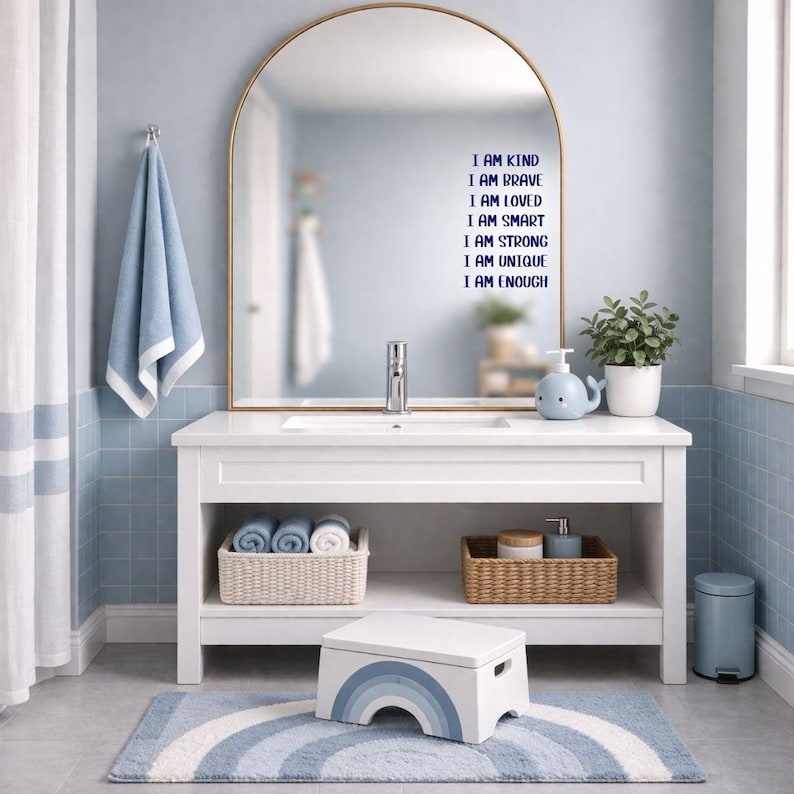 Puede incluir: Un tocador de ba&ntilde;o blanco con un espejo con marco dorado, una alfombra de arco&iacute;ris azul y blanco y un taburete. La pared sobre el tocador tiene el texto "I AM KIND, I AM BRAVE, I AM LOVED, I AM SMART, I AM STRONG, I AM UNIQUE, I AM ENOUGH."