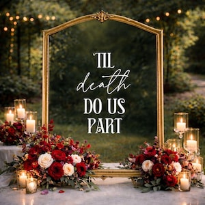 May include: A large, arched gold-framed mirror featuring the white text 'TIL death DO US PART' centered on the glass. The mirror is decorated with arrangements of deep red and white roses, greenery, and several small glass candle holders with lit candles.