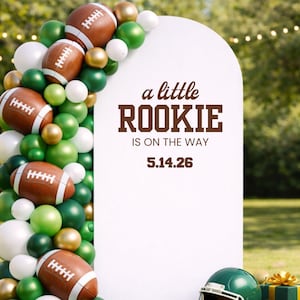 May include: A white sign with the text "a little ROOKIE IS ON THE WAY 5.14.26" is surrounded by a balloon arch of green, white, and gold balloons and footballs. A green football helmet and gift box are in the foreground.