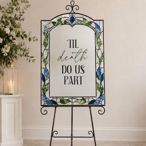 May include: A rectangular mirror with a decorative metal frame featuring stained glass accents in blue and green leaf patterns. The center of the mirror displays the black text 'TIL death DO US PART'. The mirror is supported by a metal easel stand.