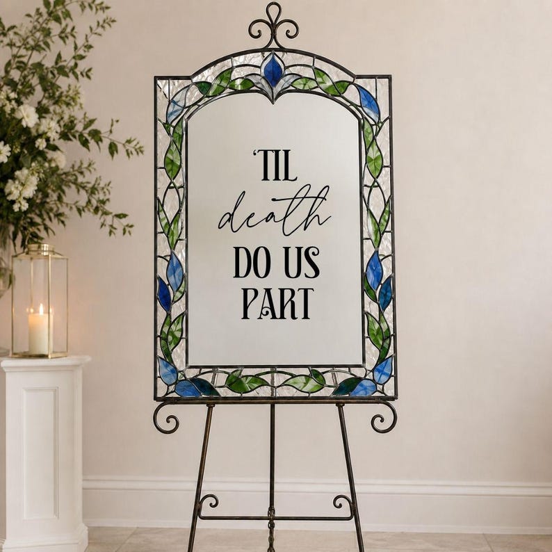 May include: A rectangular mirror with a decorative metal frame featuring stained glass accents in blue and green leaf patterns. The center of the mirror displays the black text 'TIL death DO US PART'. The mirror is supported by a metal easel stand.