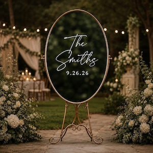 May include: An oval-shaped black sign with a gold metal frame and stand. The sign features white cursive text that reads The Smiths and the date 9.26.26. The structure is supported by a decorative gold metal base with curved legs.