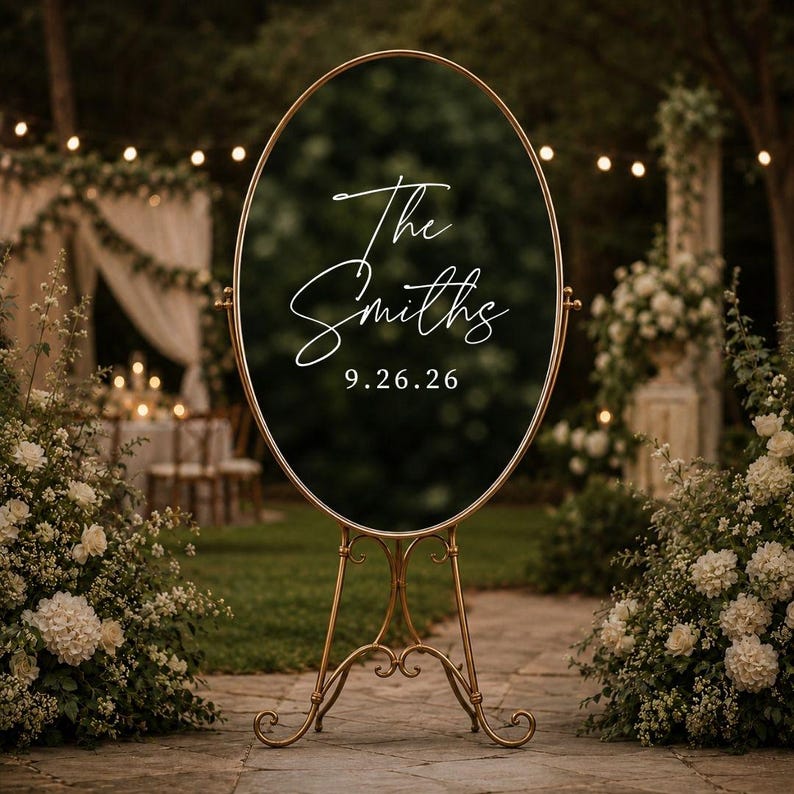 May include: An oval-shaped black sign with a gold metal frame and stand. The sign features white cursive text that reads The Smiths and the date 9.26.26. The structure is supported by a decorative gold metal base with curved legs.
