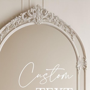 Pu&ograve; includere: Uno specchio ad arco con una cornice color crema decorata con intricati motivi a volute e intagli floreali. La superficie dello specchio riflette una parete liscia e mostra il testo bianco Custom TEXT in un mix di corsivo e stampatello.