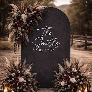 May include: A large, matte black arched sign featuring the white text "The Smiths" and the date "05.17.26". The sign is decorated with arrangements of dried pampas grass, dark red roses, and white flowers, including a crescent moon accent.