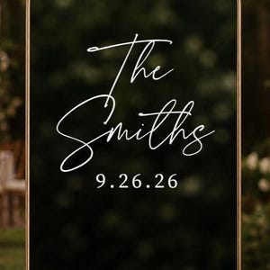 May include: An arched black sign with a thin gold metal frame. The text "The Smiths" is written in a white cursive script, and the date "9.26.26" is printed below in a white serif font. The surface is reflective and smooth.