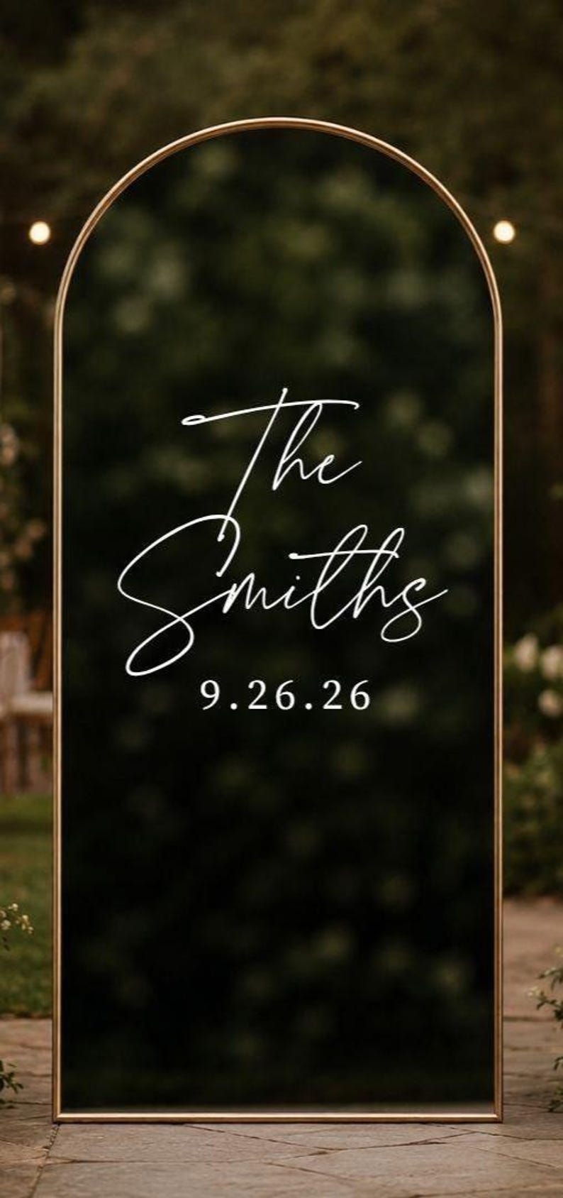 May include: An arched black sign with a thin gold metal frame. The text "The Smiths" is written in a white cursive script, and the date "9.26.26" is printed below in a white serif font. The surface is reflective and smooth.