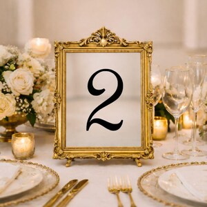 May include: A rectangular table number sign featuring the black numeral 2 centered on a white background, encased in an ornate gold-colored frame with intricate scrollwork details along the edges and a decorative crest at the top center.