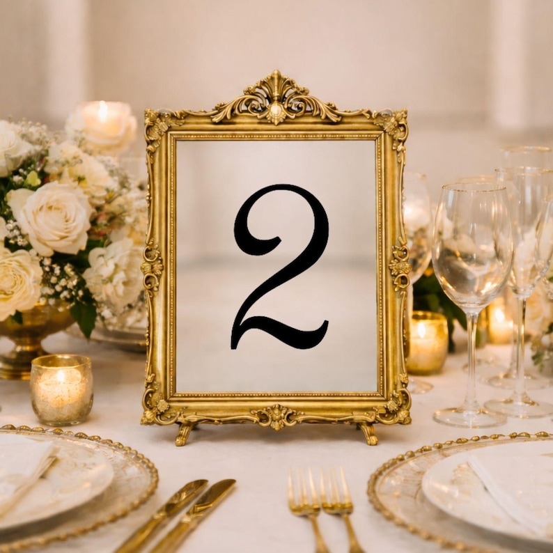 May include: A rectangular table number sign featuring the black numeral 2 centered on a white background, encased in an ornate gold-colored frame with intricate scrollwork details along the edges and a decorative crest at the top center.