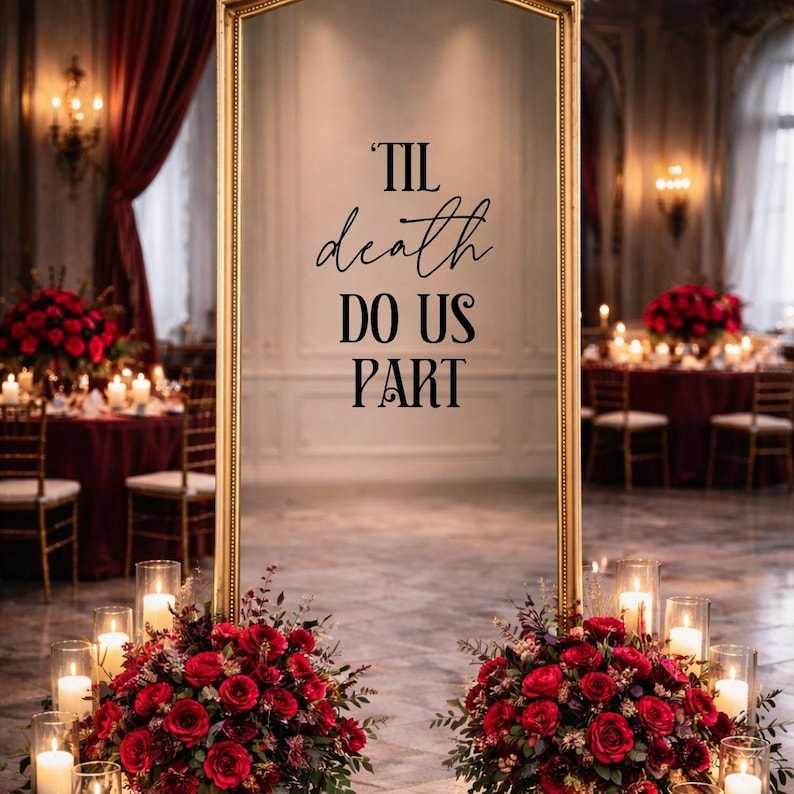 May include: A tall, rectangular floor mirror with a gold frame featuring the black text 'TIL death DO US PART' centered on the glass surface. The mirror is positioned between two large, lush arrangements of deep red roses and greenery.