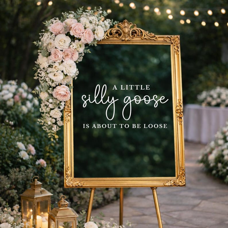 Op de afbeelding: Een goudkleurig bord op een standaard met de tekst "A little silly goose is about to be loose". Het bord is versierd met een bloemstuk van roze en witte rozen. Twee gouden lantaarns staan op de voorgrond.