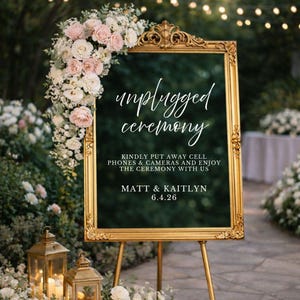 May include: A gold ornate frame holding a dark sign that reads unplugged ceremony, kindly put away cell phones & cameras and enjoy the ceremony with us, Matt & Kaitlyn, 6.4.26. A large arrangement of white and pink roses decorates the top left corner.