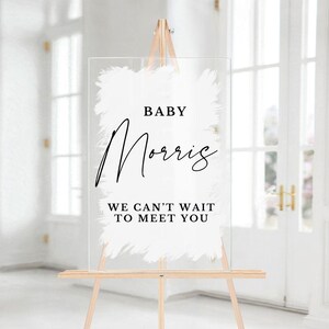 May include: White acrylic sign with a brushstroke design and black text that reads "Baby Morris We Can't Wait To Meet You".