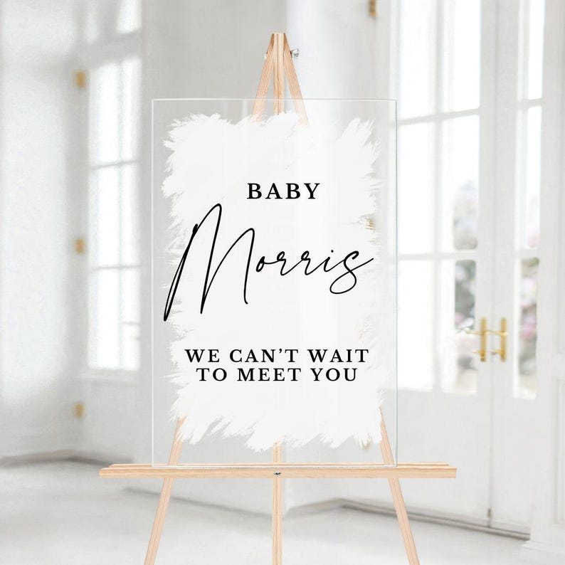 May include: White acrylic sign with a brushstroke design and black text that reads "Baby Morris We Can't Wait To Meet You".