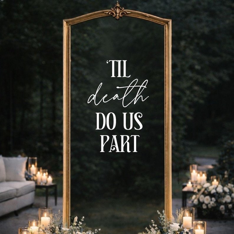 May include: A tall, rectangular mirror with an ornate gold frame featuring a decorative top crest. The glass surface displays the white text 'TIL death DO US PART' in a combination of serif and script fonts, centered within the frame.