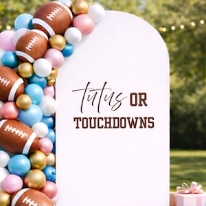 May include: A white sign with the text "Tutus OR Touchdowns" in brown script and block letters. The sign is decorated with a balloon arch of footballs, and gold, pink, white, and blue balloons. The background is a blurred outdoor scene.