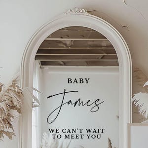 May include: A white arched mirror with a black inscription that reads "BABY James WE CAN'T WAIT TO MEET YOU".