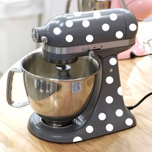May include: A gray stand mixer with white polka dots. The mixer has a silver bowl and a silver handle. The mixer is on a wooden surface.