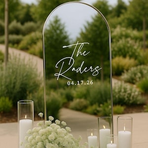 May include: An arched mirror sign with a thin gold frame featuring white cursive text that reads The Raders and the date 04.17.26. The sign is surrounded by white pillar candles in glass cylinders and a cluster of small white flowers.