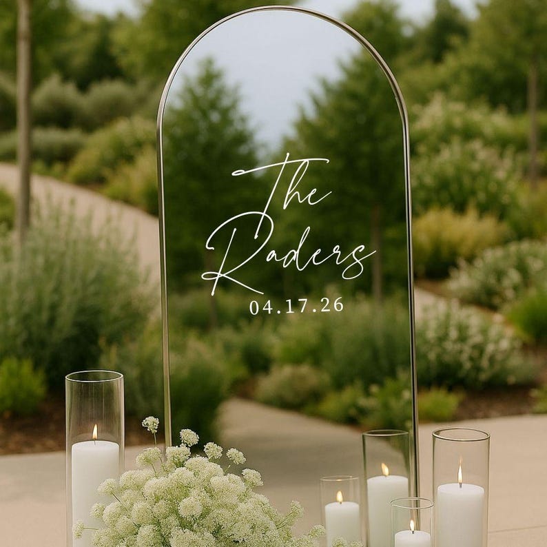 May include: An arched mirror sign with a thin gold frame featuring white cursive text that reads The Raders and the date 04.17.26. The sign is surrounded by white pillar candles in glass cylinders and a cluster of small white flowers.