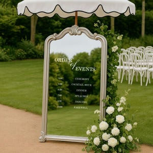 Pode incluir: Um espelho alto com moldura prateada arqueada exibindo um cronograma de eventos em texto branco, posicionado sob um guarda-sol branco com bordas recortadas. O espelho &eacute; decorado com arranjos florais brancos no lado direito.