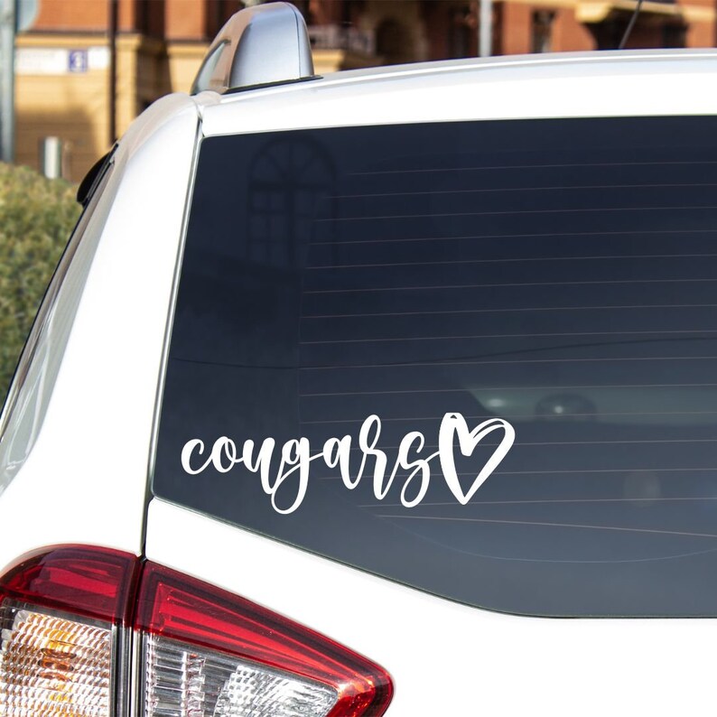 May include: White vinyl decal with the word "cougars" in cursive lettering and a heart symbol. The decal is on the back window of a white car.