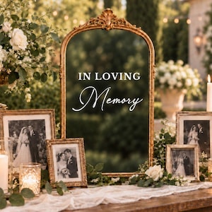 May include: An arched gold-toned mirror featuring the white text IN LOVING Memory centered on the glass. The mirror is surrounded by three framed black and white wedding photographs, white floral arrangements, and two lit glass candle holders.