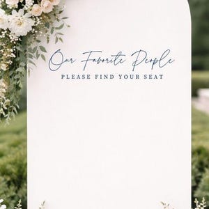 Op de afbeelding: Een wit boogvormig bord met de tekst "Our Favorite People" in een donkerblauw handschrift en "PLEASE FIND YOUR SEAT" in een donkerblauw schreeflettertype. Het bord is versierd met witte bloemen en groen blad aan de hoeken.