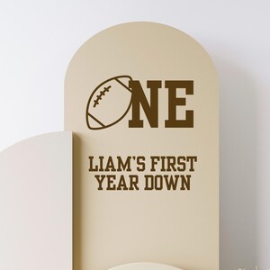 May include: A tan-colored sign with a football graphic and the word "ONE" in brown. Below, the text reads "LIAM'S FIRST YEAR DOWN" in brown. The sign has an arched top.