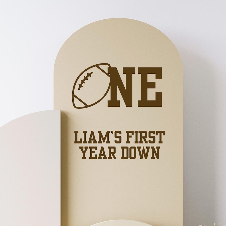 May include: A tan-colored sign with a football graphic and the word "ONE" in brown. Below, the text reads "LIAM'S FIRST YEAR DOWN" in brown. The sign has an arched top.