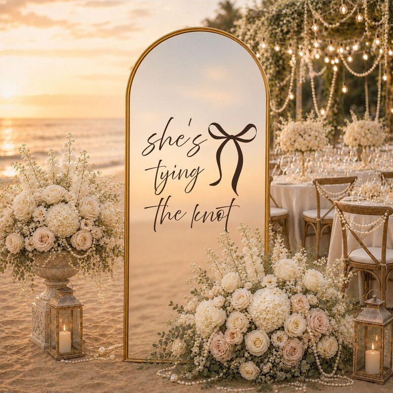 May include: An arched gold-framed mirror featuring the black text "she's tying the knot" with a bow graphic. The mirror is positioned on a sandy beach, flanked by two large white floral arrangements in stone urns and two glass lanterns.