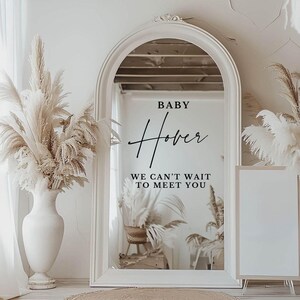 May include: A white framed mirror with an arched top, reflecting a room with a wicker chair and plants. The mirror has a black vinyl decal that reads "BABY Hover WE CAN'T WAIT TO MEET YOU".