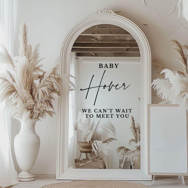 May include: A white framed mirror with an arched top, reflecting a room with a wicker chair and plants. The mirror has a black vinyl decal that reads "BABY Hover WE CAN'T WAIT TO MEET YOU".