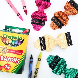 May include: A box of 24 Crayola crayons with several colorful glitter bow hair clips. The box is yellow and green with the Crayola logo and text. Several crayons are scattered around the box and hair clips.