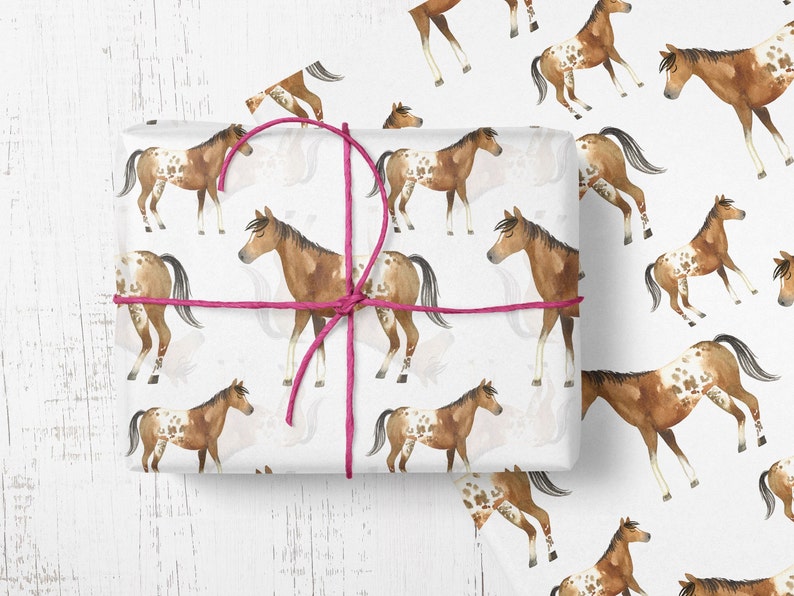 May include: White wrapping paper with a repeating pattern of brown and white spotted horses. The horses are in various poses, some are running, some are standing, and some are looking to the side. The paper is tied with a pink ribbon.