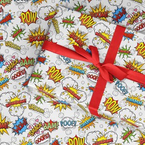 Op de afbeelding: Wit inpakpapier met een rood lint. Het papier is bedekt met een stripboek-stijl patroon met woorden als "POW", "BOOM", "KABOOM" en "SMACK" in verschillende kleuren.