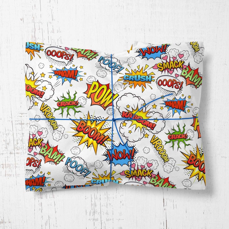 Op de afbeelding: Wit inpakpapier met een stripboekthema. Het papier is bedekt met kleurrijke spraakballonnen met woorden als "POW", "BOOM", "BAM", "CRUSH", "SMACK", "VROOOM", "WHAM", "NOW", "OOPS" en "POOF".