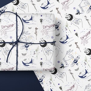 May include: White wrapping paper with a repeating pattern of hands holding crescent moons, stars, and celestial bodies. The pattern is in shades of blue, purple, and gray.