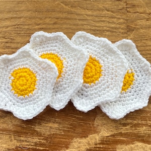 Puede incluir: Cuatro posavasos de ganchillo blancos con centros amarillos, que se asemejan a huevos fritos. Los posavasos están dispuestos en una pila, con los centros amarillos hacia afuera.