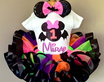 Puede incluir: Un atuendo de Halloween para un niño de un año. El conjunto incluye una camiseta blanca con un diseño de Minnie Mouse en forma de murciélago, un tutú colorido y orejas con lentejuelas y un lazo rosa. El nombre "Mira" está impreso en la camiseta.