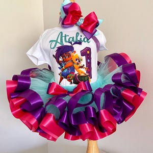 May include: A white t-shirt with the name "Atalia" and a cartoon graphic of two characters, paired with a tutu skirt. The skirt features layers of purple, pink, and teal ribbons. A large bow with matching colors sits atop the shirt.