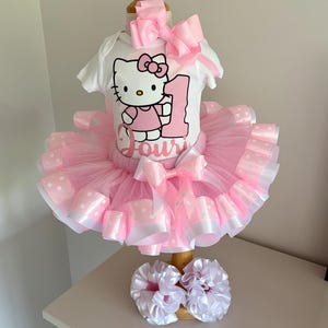 Puede incluir: Un body blanco de manga corta con un gráfico de Hello Kitty, el número 1 y el nombre Jouri. El conjunto incluye una falda de tul rosa con ribete de cinta de lunares blancos, un lazo rosa grande para el pelo y dos coleteros blancos con volantes.