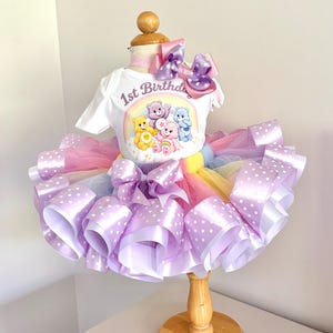 May include: A white t-shirt with a Care Bears graphic and "1st Birthday" text, paired with a multi-colored tutu skirt. The skirt features layers of tulle and lavender ribbon with white polka dots. A large bow completes the outfit.