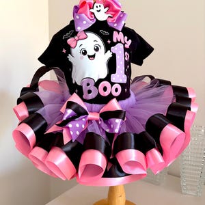 May include: A black short-sleeved shirt featuring a white ghost graphic with a pink bow and the text My 1st Boo, paired with a layered purple tulle tutu skirt trimmed with alternating black and pink satin ribbons and a matching hair bow.