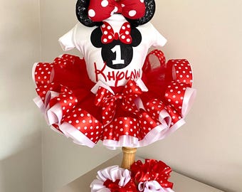 May include: A white short-sleeved shirt with a black glittery mouse silhouette, a red polka-dot bow, the number 1, and the name Khlynn in red script. It includes a red tulle tutu with polka-dot ribbon trim, mouse ears, and two hair scrunchies.