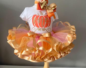 May include: A white onesie with a pumpkin design and the word "one" in gold glitter. The onesie is paired with a tulle skirt in shades of orange and pink, accented with gold satin ribbon. A matching orange and gold bow is included.