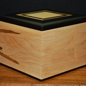 May include: A wooden box with a light-colored, natural wood grain body. The top is black with a gold-colored square inlay. The box is square-shaped and sits on a wooden surface. The box has a smooth, polished finish.