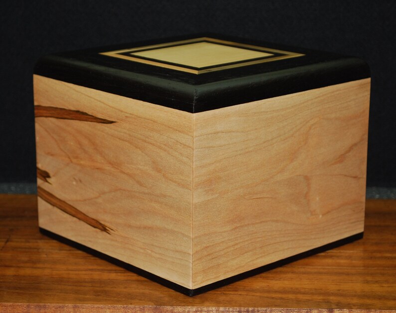 May include: A wooden box with a light-colored, natural wood grain body. The top is black with a gold-colored square inlay. The box is square-shaped and sits on a wooden surface. The box has a smooth, polished finish.