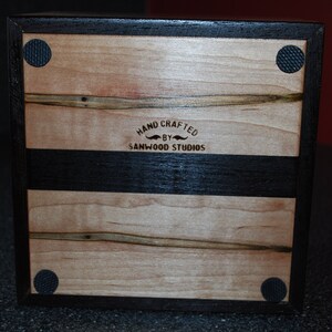 May include: A handcrafted wooden box with a light wood grain top and bottom, and a dark wood border and stripe. The text "HAND CRAFTED BY SANWOOD STUDIOS" is visible. Black rubber feet are on the corners.