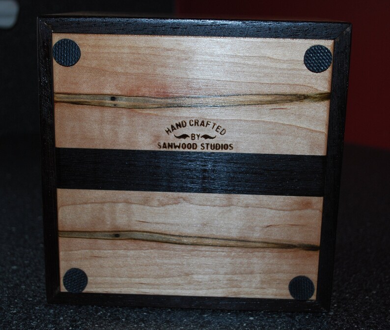May include: A handcrafted wooden box with a light wood grain top and bottom, and a dark wood border and stripe. The text "HAND CRAFTED BY SANWOOD STUDIOS" is visible. Black rubber feet are on the corners.